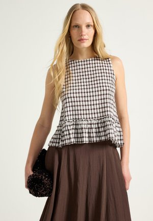 Lyshåret kvinde iført sort-hvid ternet top uden ærmer med flæsekant og lang brun nederdel, holder en sort tekstureret clutch.