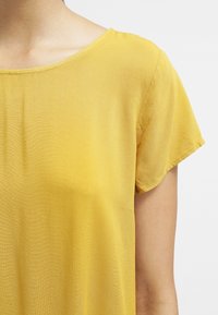 Haut jaune à manches courtes avec une coupe décontractée et un tissu léger et lisse. L'encolure est de style ras du cou avec des coutures discrètes.