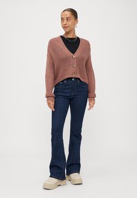 Cárdigan de canalé rosa con tres botones, llevado sobre una camiseta negra. Combinado con jeans de mezclilla oscuros de pata de elefante y zapatillas gruesas color beige.