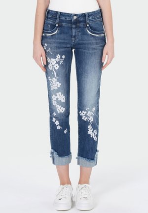 Blauwe cropped spijkerbroek met witte bloemenprint op de benen, rafelige zomen en witte sneakers gedragen door een persoon die staat tegen een effen achtergrond.