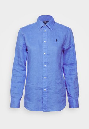Chemise longue à manches en lin bleu clair, style boutonné, avec un petit logo noir sur la poitrine gauche et des boutons blancs.