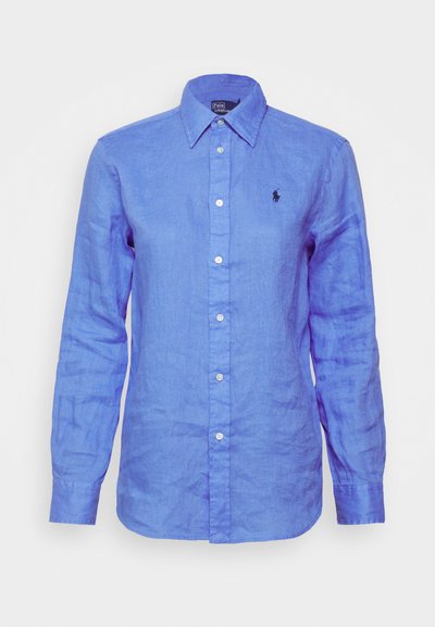 Ανοιχτό μπλε λινό πουκάμισο με μακριά μανίκια, σε στυλ button-down, που διαθέτει ένα μικρό μαύρο λογότυπο στο αριστερό στήθος και λευκά κουμπιά.