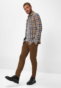 Karierte Hemd in Blau, Braun und Creme, über einem grauen Pullover getragen; braune Hose und schwarze Schuhe. Verfügt über geknöpfte Taschen und einen lockeren Schnitt.