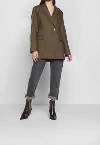 Femme portant un blazer vert olive, un jean noir foncé retroussé et des bottines marron foncé brillantes à bout pointu et petit talon.