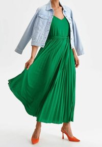 Grön plisserad maxiklänning med matchande bälte, ihop med en ljus denimjacka. Accenterad med klara orangea högklackade skor.