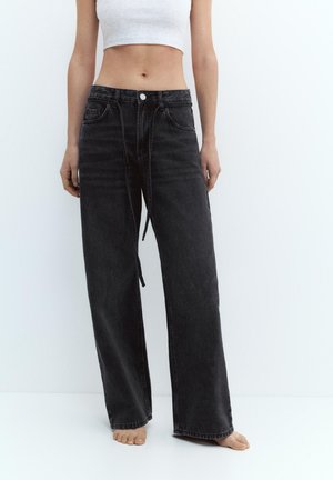 Nő magas derekú, széles szárú fekete farmert és fehér, rövid felsőt visel, mezítláb áll egyszerű, fehér háttér előtt.