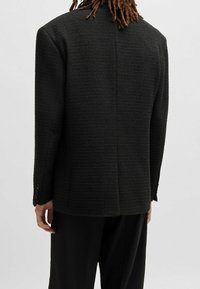 Blazer nero testurizzato con una forma strutturata, fessura centrale posteriore e polsini abbottonati, mostrato da dietro su uno sfondo semplice.