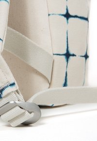 Gros plan d'une sangle en tissu beige avec boucle en métal attachée à un sac avec un motif quadrillé bleu sur fond blanc.