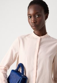 Camicia button-up rosa chiaro con colletto alla mandarino, abbinata a una piccola borsa blu con manico texturizzato.