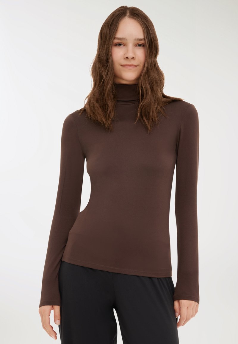 Donna con lunghi capelli castani mossi, che indossa un dolcevita marrone scuro aderente e pantaloni neri, in piedi contro uno sfondo chiaro e uniforme.