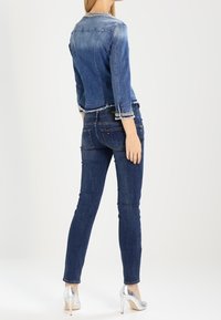 Nainen seisoo, päällä tiukasti istuva denim-takki, jossa on pitsireunus, tummat skinny-farkut ja hopeiset korkokengät, nähtynä takaa.