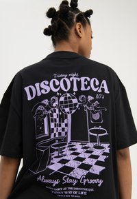 Persoon met bantu-knotkapsel draagt een zwart t-shirt met een paars jaren 80-discothema met cherubs, discobal, dambordvloer en tekst.