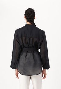 Cămașă neagră, semi-transparentă, cu mâneci lungi, manșete cu butoane și talie împăturită. Partea din spate are un design plisat și un tiv rotunjit.
