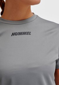 Szara sportowa koszulka z krótkim rękawem, okrągłym dekoltem i teksturowaną tkaniną. Na piersi nadrukowane logo "HUMMEL" w grubym czarnym kolorze.