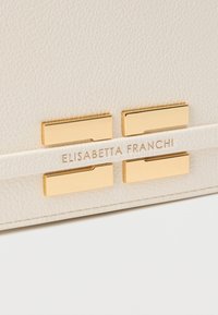 Λευκή τσάντα από δερμα με υφή, εξοπλισμένη με χρυσούς μεταλλικούς τόνος και γεωμετρικά διακοσμητικά, με το όνομα της μάρκας "ELISABETTA FRANCHI" ανάγλυφο.