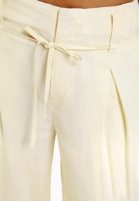 Krämfärgade, löst sittande byxor med knytmidja, framvikt och sidofickor. Tyget verkar vara lätt och mjukt.