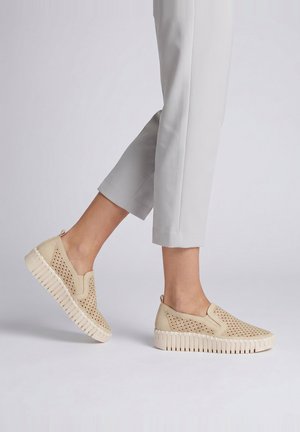 Person iført beige slip-on sneakers med perforeret detalje og tykke hvide såler, kombineret med lysegrå cropped bukser.
