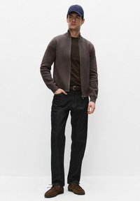 Man wearing blue cap, brown zip-up sweater, dark green shirt, black jeans, and brown shoes standing with one hand in pocket.