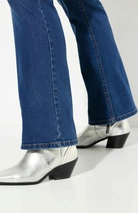 Hopeat nahkasaappaat, joissa on terävä kärki ja matala musta korko, yhdistettynä tumma sinisiin laineileviin farkkuihin, joissa näkyy ompeluyksityiskohtia helmaosassa.