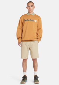 Sudadera naranja con el logo de "Timberland", camisa verde de cuello por debajo, pantalones cortos beiges, calcetines blancos y zapatillas marrones con acentos azules.