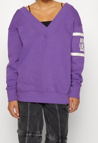 Lila oversized sweatshirt med djup V-urringning, ribbade ärmslut och en grafisk logotyp på vänster ärm. Mjukt bomullsmaterial, minimalistisk design.