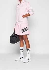 Světle růžová mikina na zip s bílými pruhy, odpovídající růžové šortky s černými pruhy, černá taška, bílé tenisky a pruhované bílé ponožky.