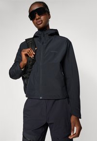 Person wearing black hooded jacket, matching pants, large black sunglasses, holding a black backpack strap over one shoulder against neutral background.