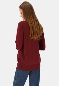 Bordowy sweter z długim rękawem o zaokrąglonym dolnym brzegu, miękkiej fakturze i prążkowanych detalach, w połączeniu z niebieskimi dżinsami.