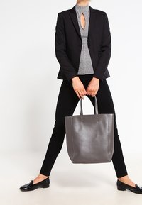 Grå läder tote-väska med två robusta handtag, slät textur och kontrasterande söm. Bärs av en person i figurnära svarta kläder.