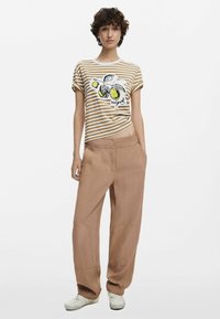 Randig beige och vit t-shirt med en citron-illustration, kombinerad med lösa bruna byxor med en slät yta och synliga sömmar. Vita skor.