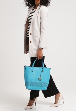 Femme en blazer blanc, chemise rayée, pantalon noir et talons, tenant un sac cabas turquoise avec des détails perforés et un pendentif.