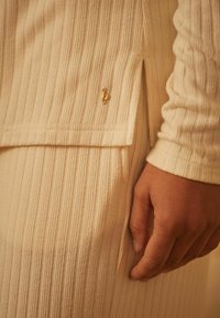 Beige geribbelde breisel met een lichte zijsplit en een gouden metalen logo-accent nabij de zoom. Close-up van een hand die op de stof rust.