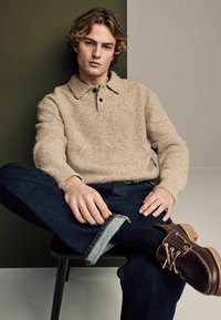 Beež tekstuuriga kudum polo-sweater, tumedad teksapüksid üleskeeratud äärega ja pruunid lipsuga saapad heledate nööpidega, istumisposeeringus toolil.