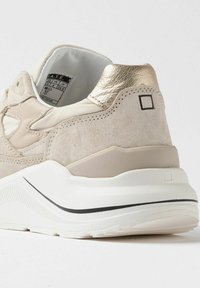 Sneaker beige con un upper in suede testurizzato, dettaglio in oro metallizzato sul tallone e una suola bianca chunky con una striscia nera sul lato.