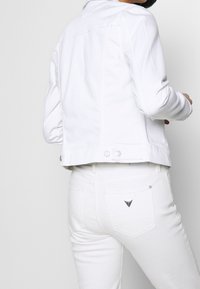 Nainen, jolla on päällään valkoinen denimi takki ja valkoiset farkut, joissa on kolmiomainen metallilogo takataskussa, tasaisella taustalla.