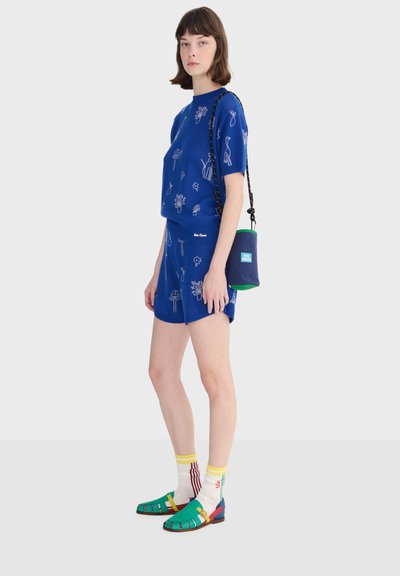 Femme portant un haut bleu à motifs et un short, avec un petit sac bandoulière bleu et vert, des chaussettes blanches et des sandales colorées.