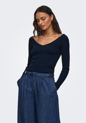 Žena s dlouhými tmavými vlasy, která má na sobě námořnicky modrý top s výstřihem do V a dlouhými rukávy a vysokým pasem široké modré džíny s rukama v kapsách.