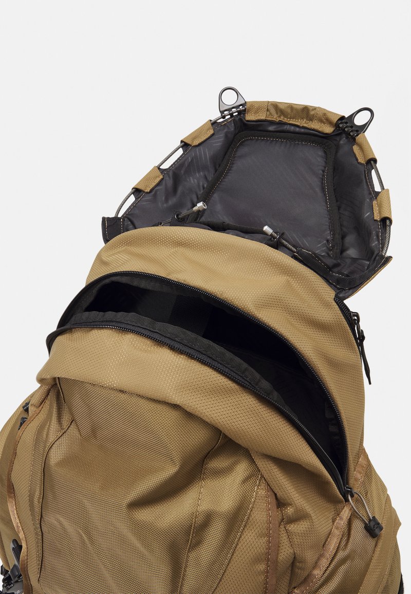 Zaino color sabbia con un esterno testurizzato, dotato di un compartimento principale con zip e una parte superiore aperta resistente con hardware regolabile.