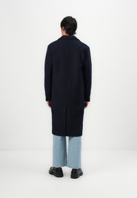 Cappotto di lana blu navy con taglio dritto, collo a scendenti e maniche lunghe. Indossato su jeans svasati azzurri e calzature nere.