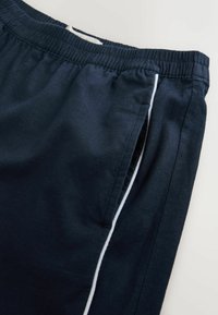 Pantalon à taille élastique bleu marine avec un détail de passepoil blanc sur le côté et une poche latérale sur un fond clair.