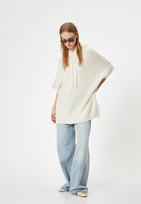 Suéter grande de punto en color crema con textura de canalé y capucha, combinado con jeans de pierna ancha en azul claro y zapato tipo slip-on en color nude.