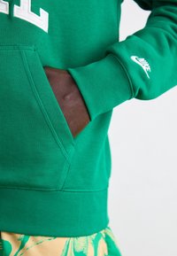 Grön bomullshoodie med känguruficka, ribbad mudd och vit Nike-logga på ärmen. Mjuk textur, avslappnad design.