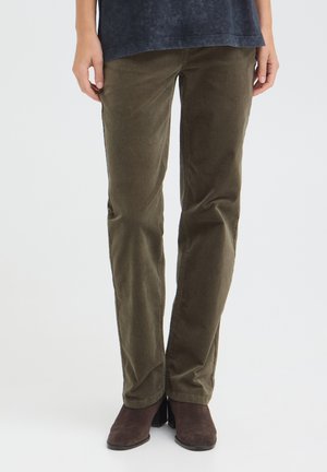 Personne portant un pantalon en velours côtelé vert olive et des bottines marron foncé, debout devant un fond blanc uni.