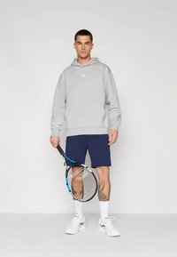 Γκρι φούτερ με κουκούλα, σκούρα μπλε σορτς, λευκά αθλητικά παπούτσια. Το μοντέλο κρατά ρακέτα τένις, με τατουάζ στο κάτω μέρος του ποδιού. Απλό φόντο.