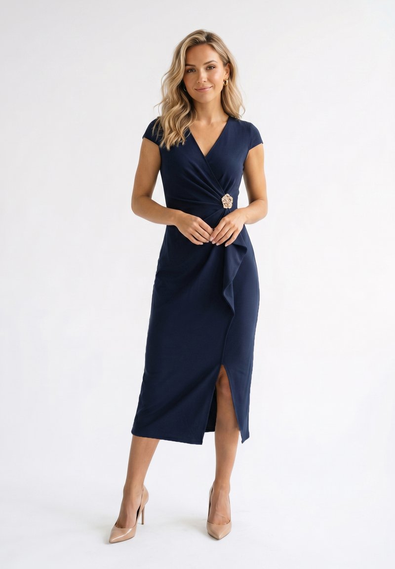 Femme en robe portefeuille bleu marine avec fente latérale et talons hauts beiges, debout sur fond blanc uni.