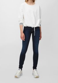 Sudadera blanca con cordón en el bajo y puños a rayas, combinada con jeans skinny oscuros y zapatillas blancas con acentos plateados.