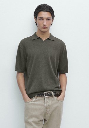 Junger Mann mit dunklen Haaren, der ein olivgrünes kurzärmliges Poloshirt und beige Hosen mit geflochtenem Gürtel trägt, die Hände in den Taschen, neutraler Hintergrund.