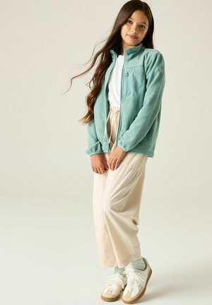 Ragazza giovane con capelli lunghi che indossa una giacca verde chiaro con cerniera, camicia bianca, pantaloni beige a gamba larga, calzini a righe e scarpe da ginnastica bianche.