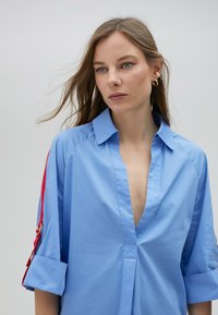 Camicia blu con bottoni, scollo a V profondo, maniche arrotolate e accenti rossi sulle spalle. Realizzata in un tessuto morbido e leggero.