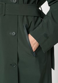 Manteau vert foncé avec ceinture, boutons, une poche zippée avec une main à l'intérieur, et un logo brodé sur le poignet de la manche.